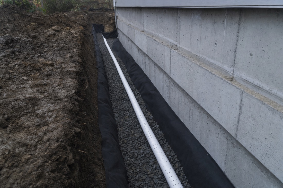 Excavated trench along a house foundation with new perforated PVC perimeter drain pipe on clean drainage gravel in the Fraser Valley